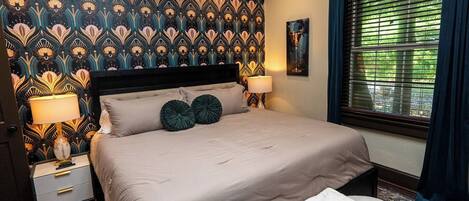 A bedroom with a patterned wallpaper, a bed with two pillows, bedside lamps, a window with blinds, and a rug.
