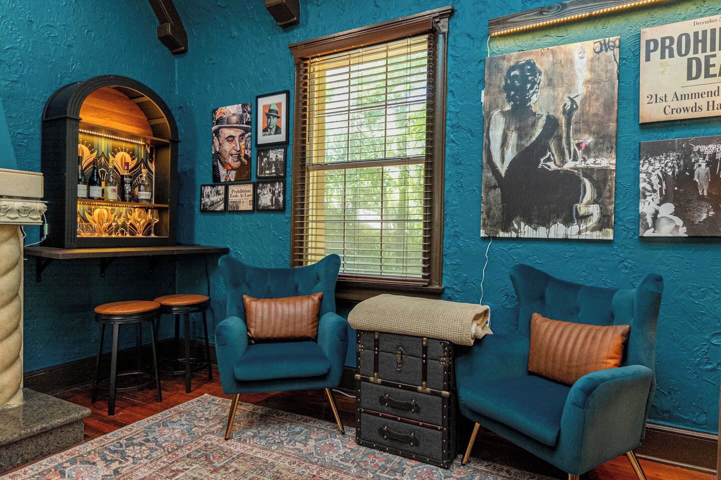 A room with teal walls, two blue armchairs with brown cushions, a bar area with bottles and glasses, and a window with blinds.