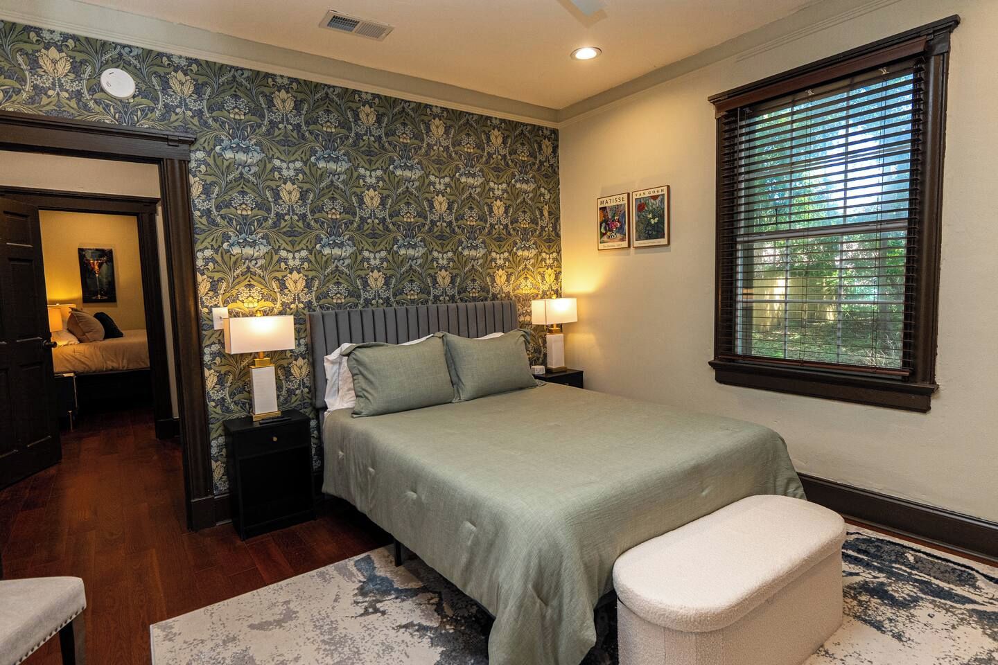 A bedroom with a bed, nightstands, a window with blinds, and a door leading to another room.