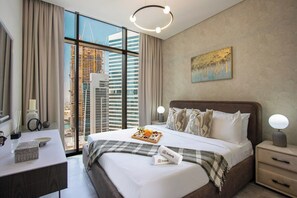 A modern hotel room with a large bed, a bedside table, a desk, and a view of the cityscape through large windows. - New Waterfront 1B with Skyline & Canal View (Dubai)