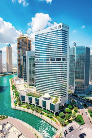 A modern high-rise building with a glass facade, surrounded by a water body and other high-rise buildings. - New Waterfront 1B with Skyline & Canal View (Dubai)