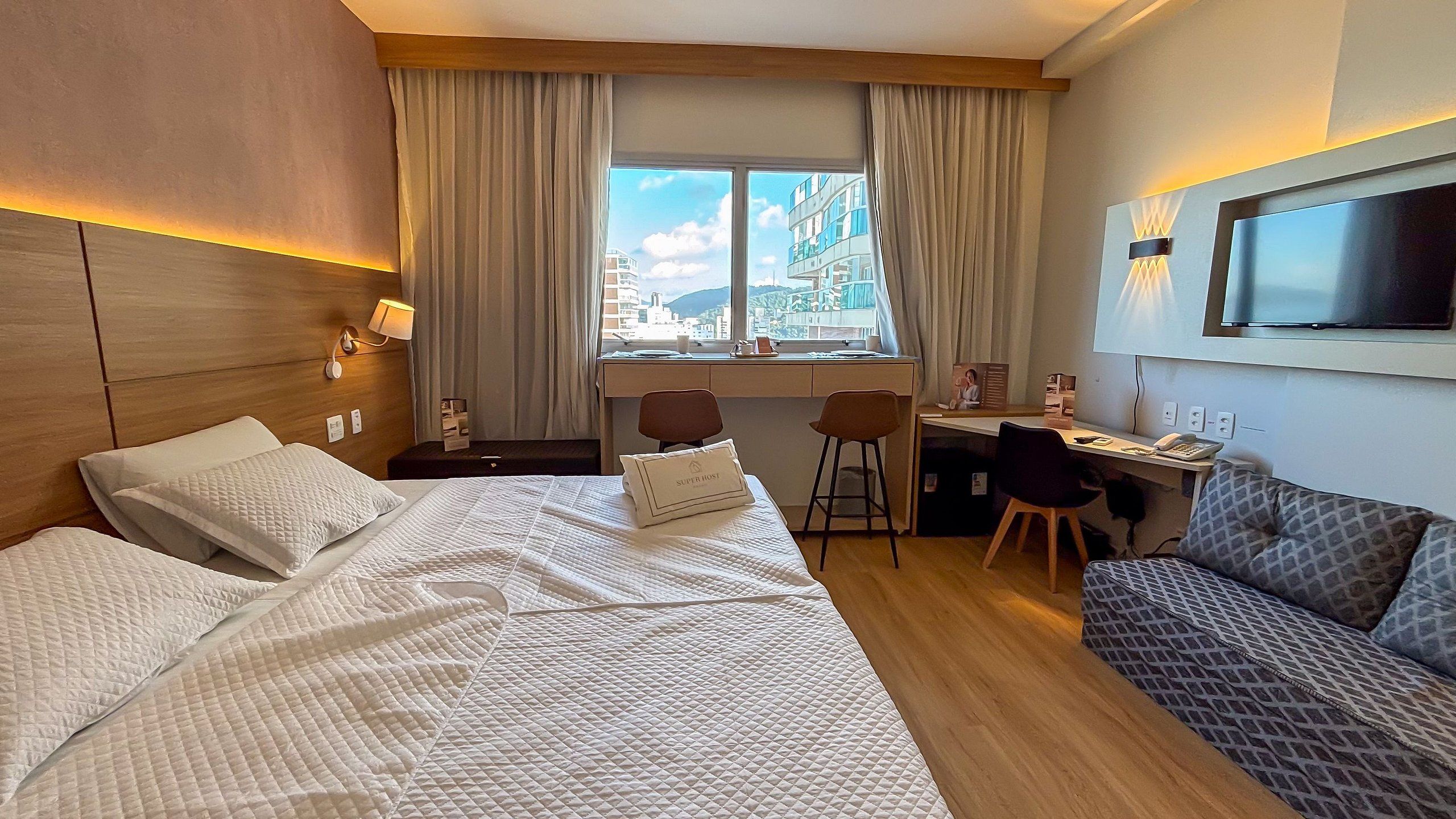 A hotel room with a bed, desk, chairs, a TV, and a window with a city view.