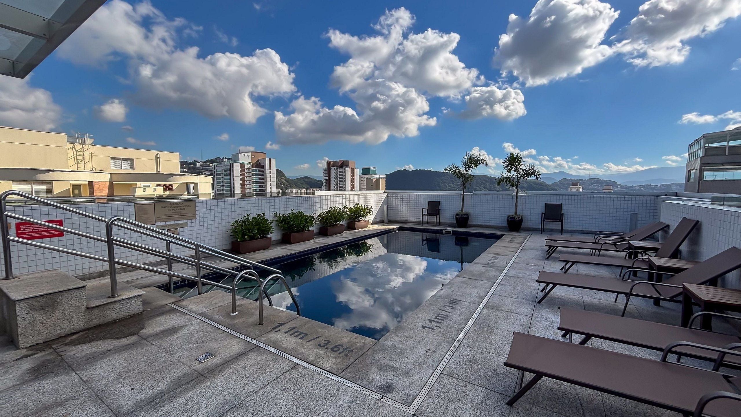 A rooftop pool with lounge chairs, a small pool area, and a view of surrounding buildings and mountains.