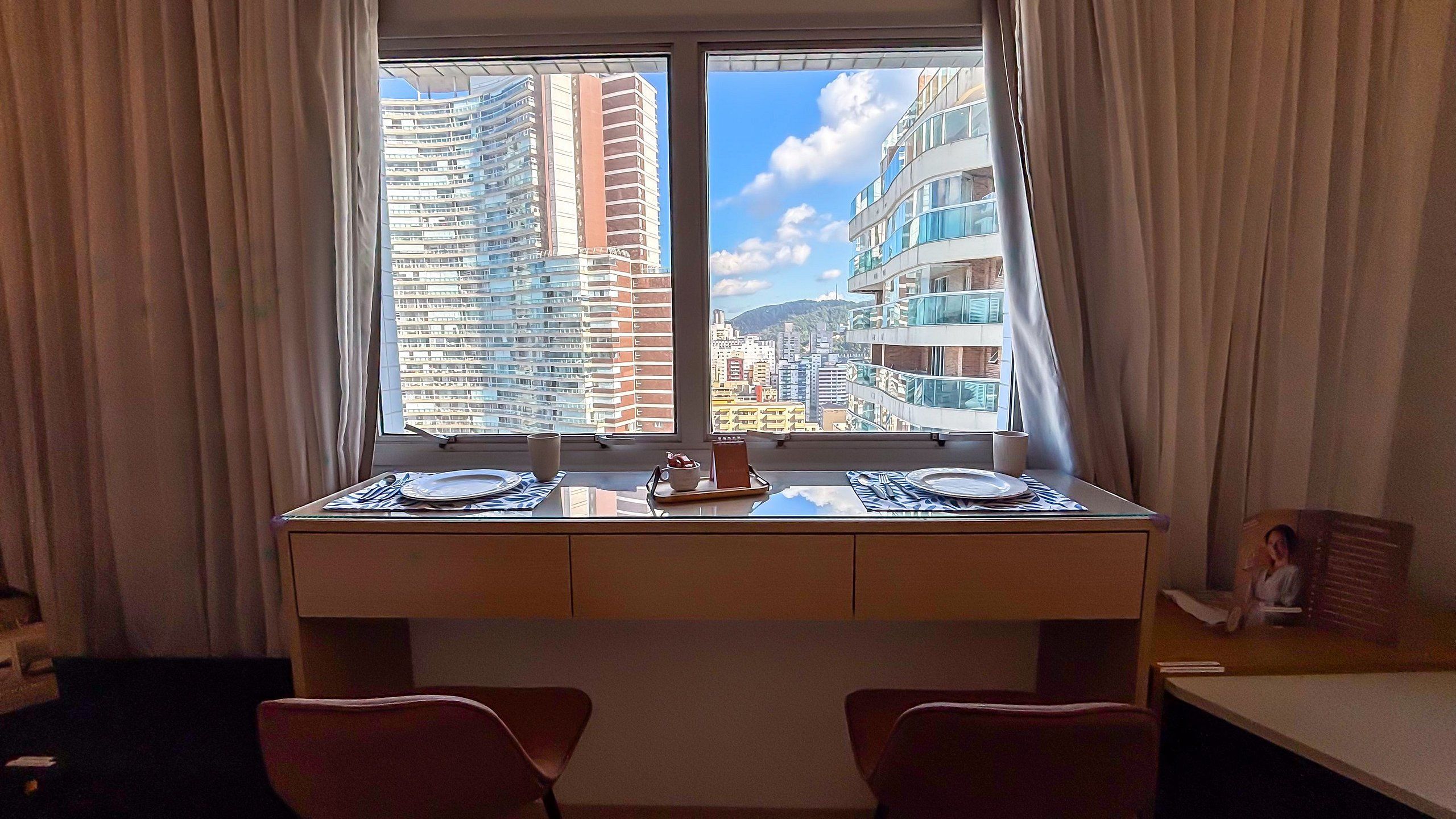 Habitación de hotel con una mesa de comedor dispuesta para dos personas, vistas a rascacielos y una ventana con cortinas.