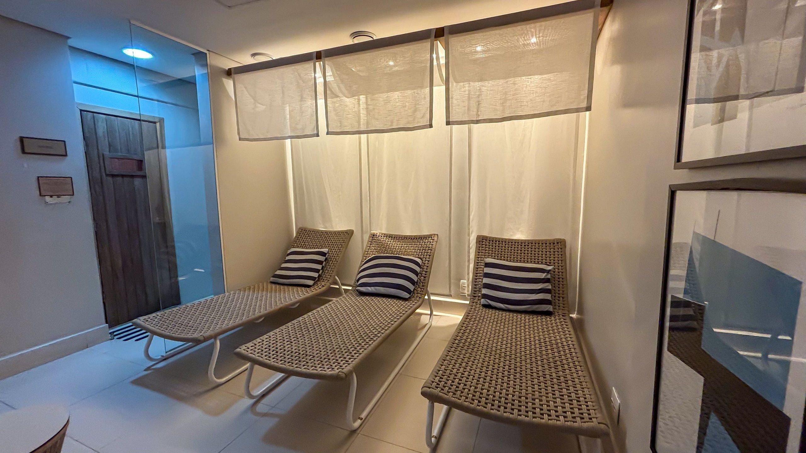 Habitación con tres sillones de mimbre, cada uno con un cojín a rayas, y un ventanal con cortinas translúcidas.