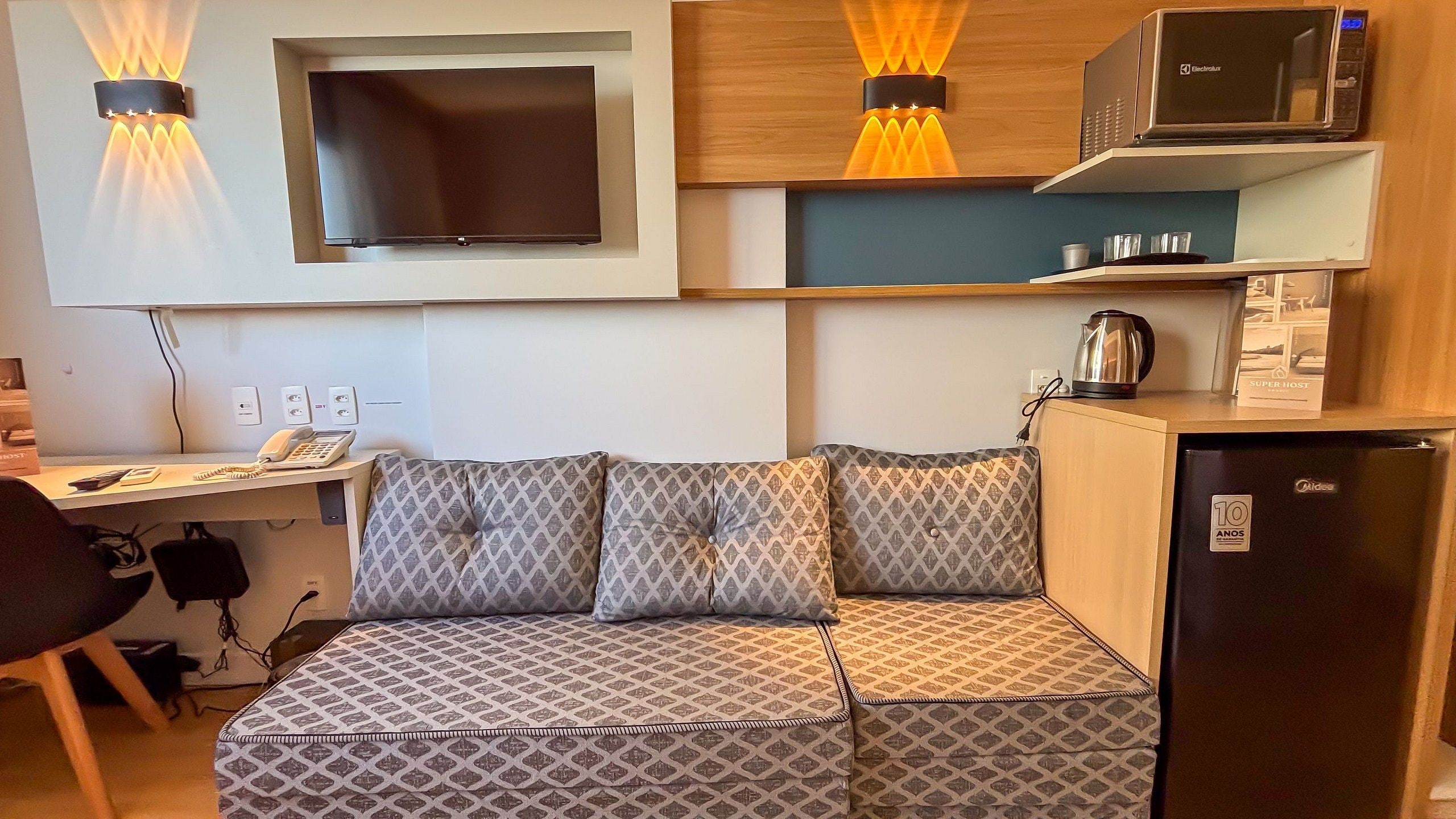 A compact kitchenette with a microwave, kettle, and a refrigerator. There is a sofa with patterned cushions and a flat-screen TV mounted on the wall.