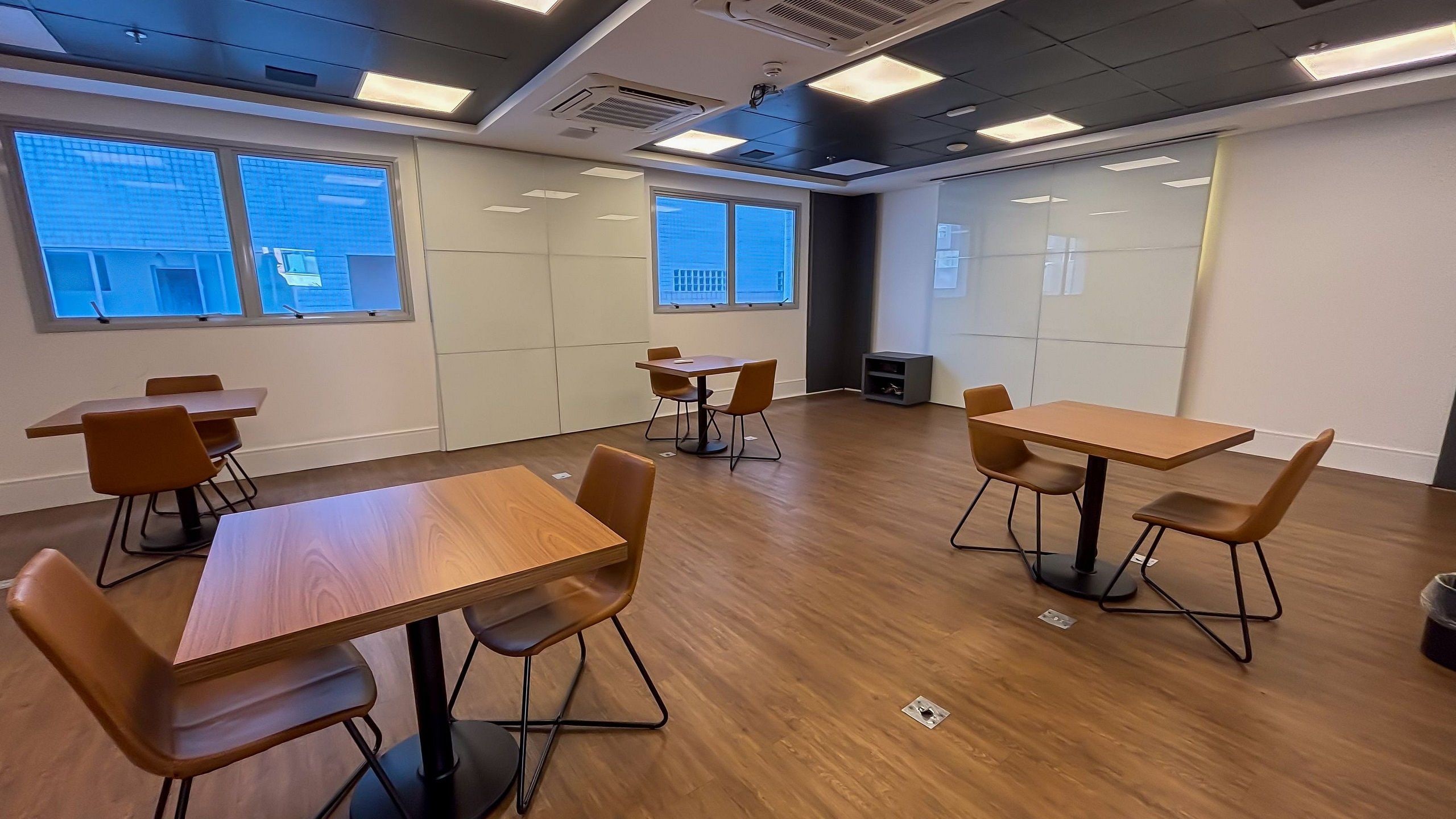 Uma sala de conferências com mesas e cadeiras de madeira, janelas amplas com persianas e um teto moderno com iluminação embutida.