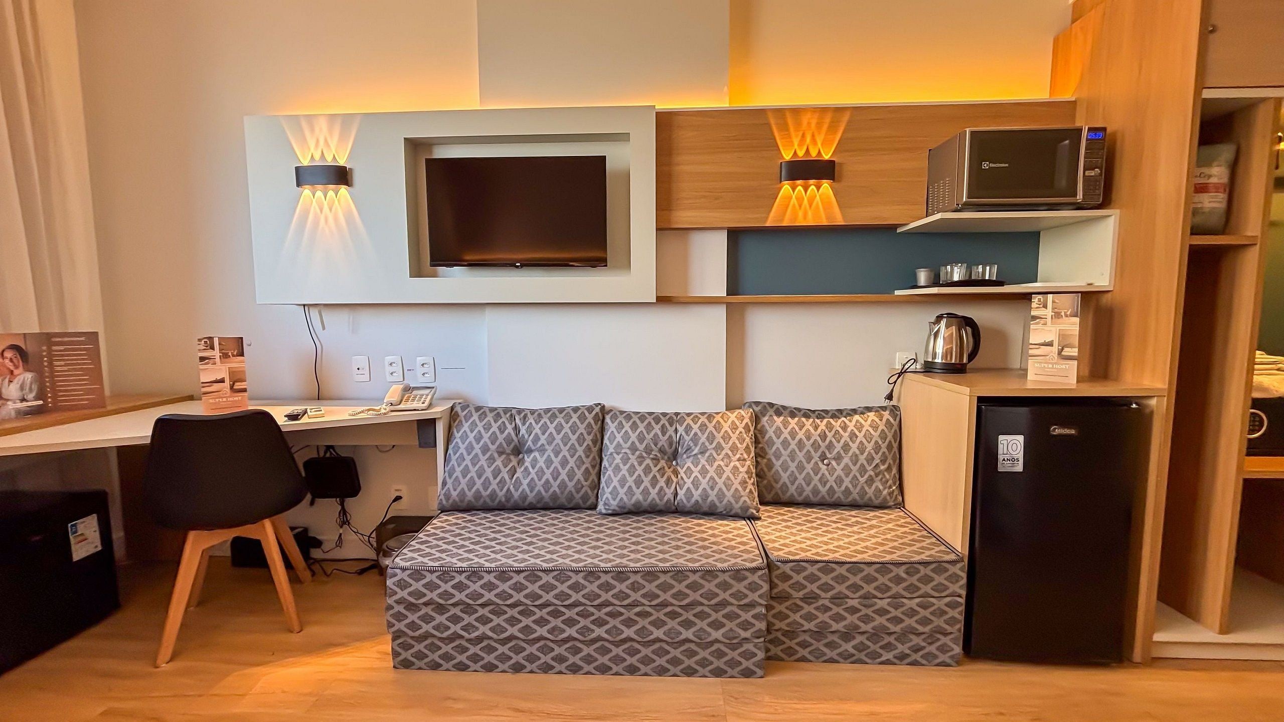 A compact hotel room with a sofa, a desk, a microwave, a refrigerator, and a wall-mounted TV.