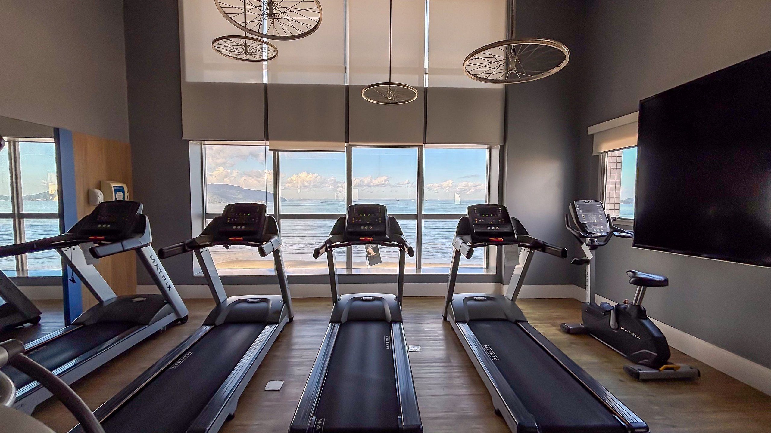 Un gimnasio con cintas de correr, una bicicleta estática y una máquina de remo. Amplias ventanas ofrecen vistas al mar.