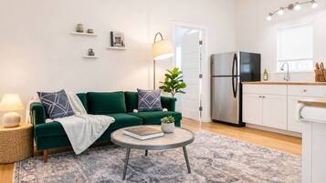 Ein modernes Wohnzimmer mit einem grünen Sofa, einem runden Couchtisch und einer Küchenzeile inklusive Kühlschrank und Schränken.