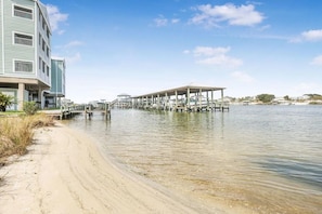 Een pand aan het water met een overdekte steiger die in het water uitsteekt, een zandstrand en een meerlaags gebouw ernaast.