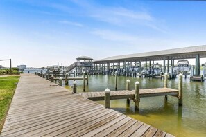 Een houten steiger die uitkomt bij een jachthaven met boten en een overdekte constructie.