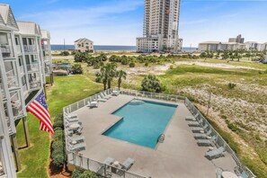A swimming pool with lounge chairs, surrounded by a fence, with a view of the ocean and buildings in the background. - Old River Landing /Jubilee Landing 306/Boaters Welcome/Outdoor Pool (Orange Beach)