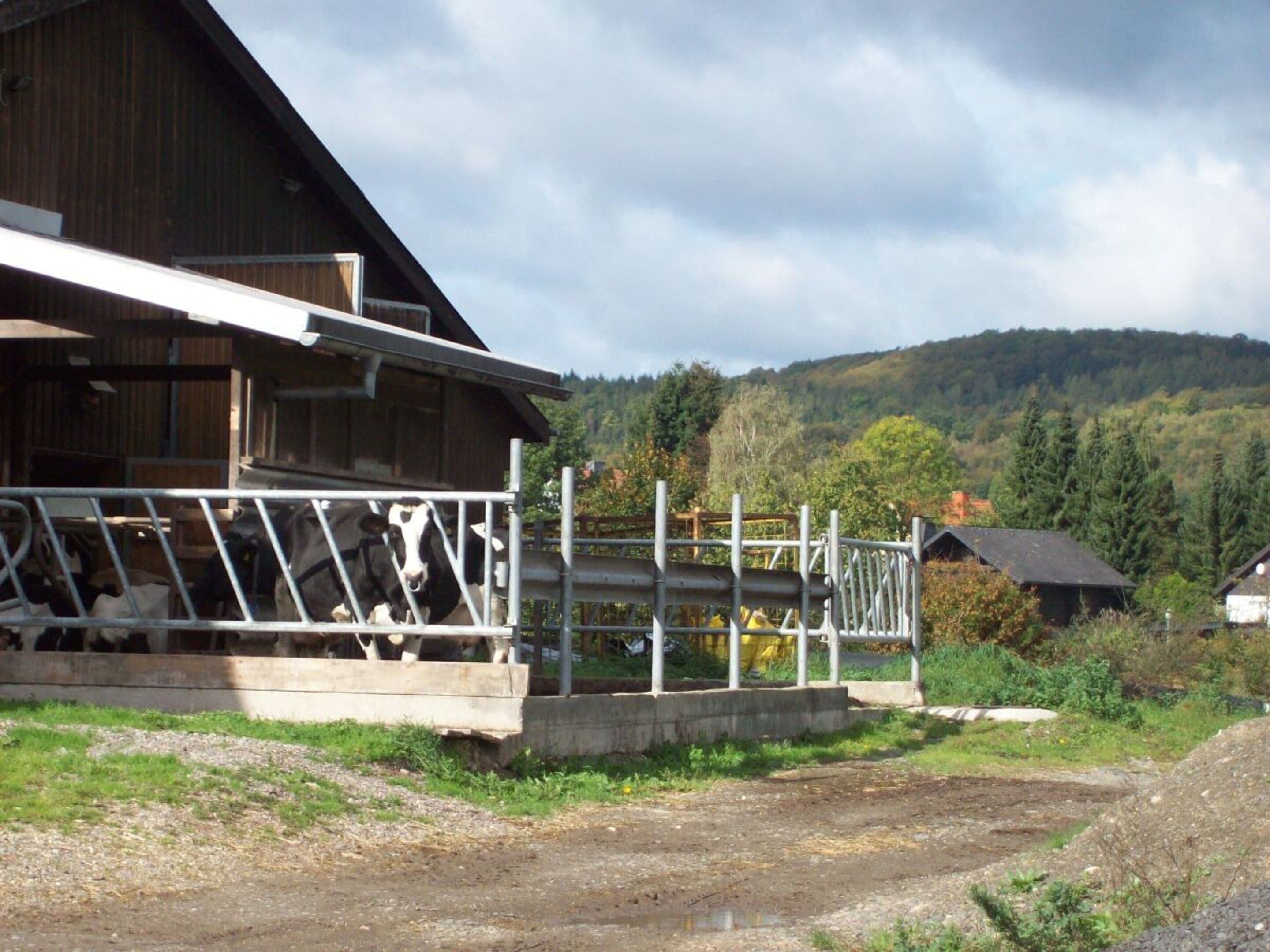 Um celeiro rústico com cerca de metal e plataforma de madeira, rodeado por uma paisagem rural com colinas e árvores.