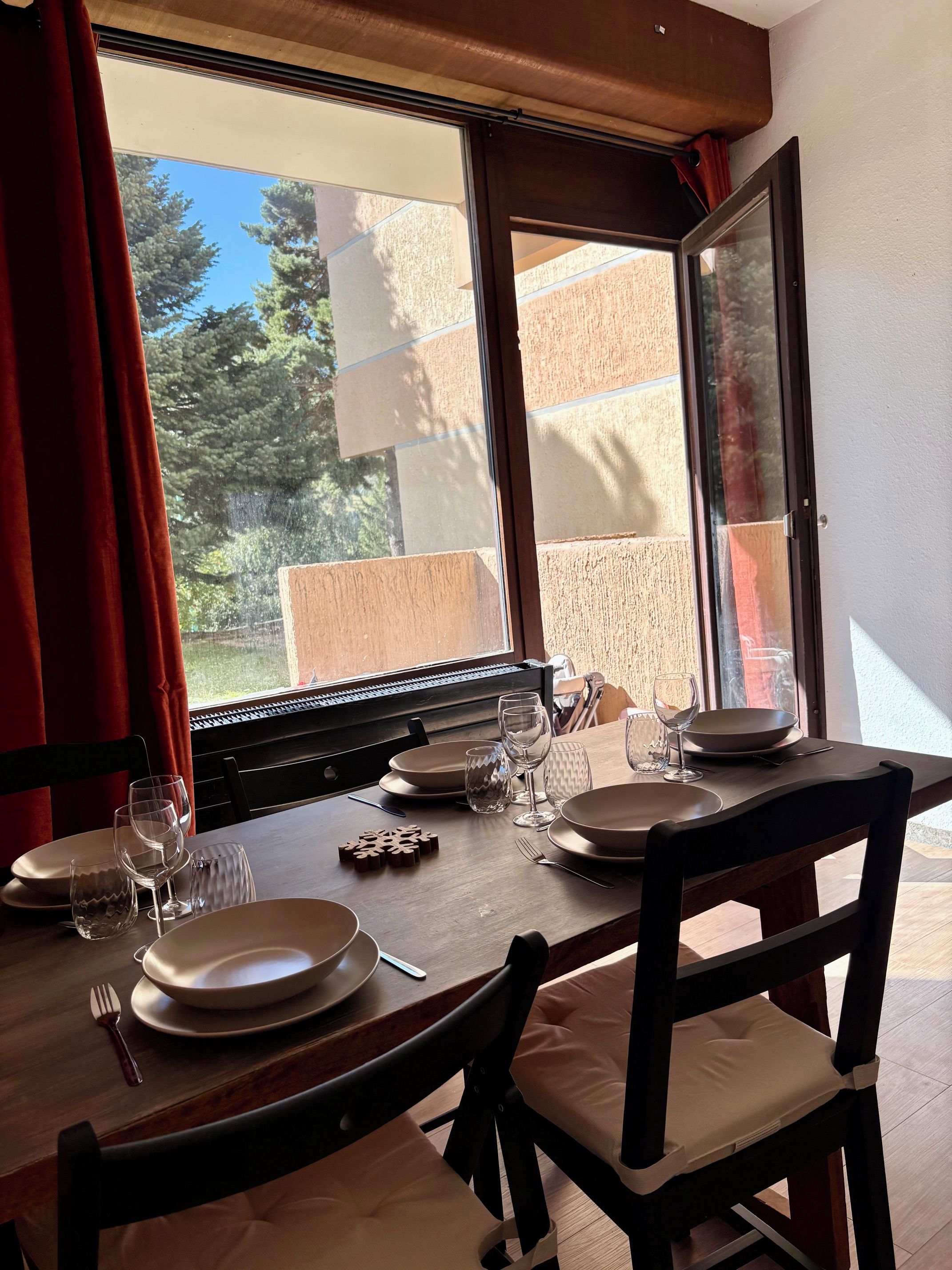 Une table à manger dressée avec des assiettes, des verres et des couverts, donnant sur des arbres et des bâtiments à travers une fenêtre.