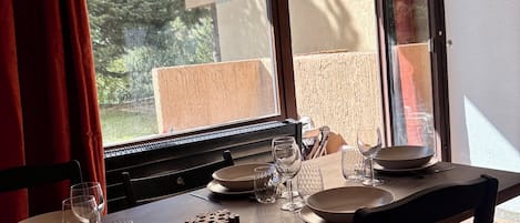 Una mesa de comedor dispuesta con platos, vasos y cubiertos, con vistas a árboles y edificios a través de una ventana.