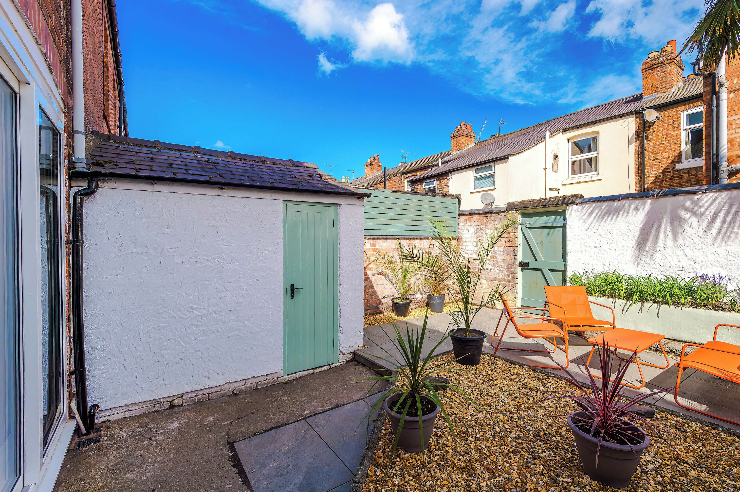 A residential courtyard with a white garage, green door, potted plants, and outdoor seating.