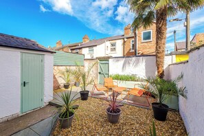 Un patio trasero residencial con una puerta verde, mobiliario exterior, plantas en macetas y un cielo azul despejado.