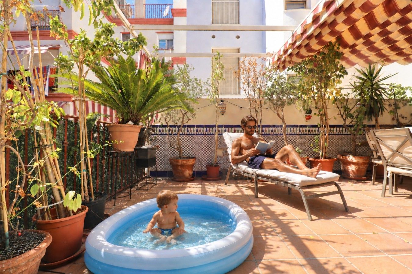 Zona de piscina con un niño jugando en una piscina inflable pequeña, un hombre leyendo un libro y varias plantas en macetas.