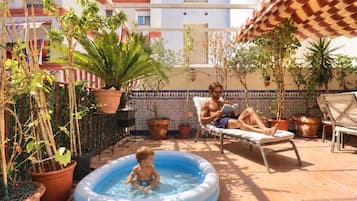 Zona de piscina con un niño jugando en una piscina inflable pequeña, un hombre leyendo un libro y varias plantas en macetas.