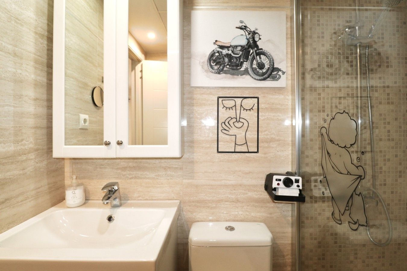 A bathroom with a sink, mirror, toilet, and a shower. There are framed pictures on the wall.