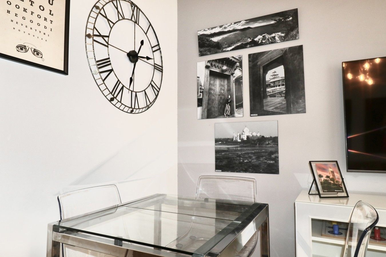 Une pièce avec une table en verre, des chaises, une horloge murale et des photographies encadrées.
