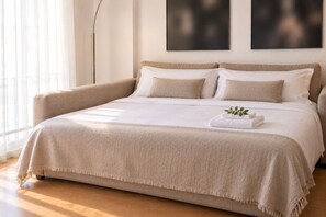Ein großes Bett mit beige farbenem Bettbezug und Kissen, ein Nachttisch mit einer Pflanze und eine Stehlampe.