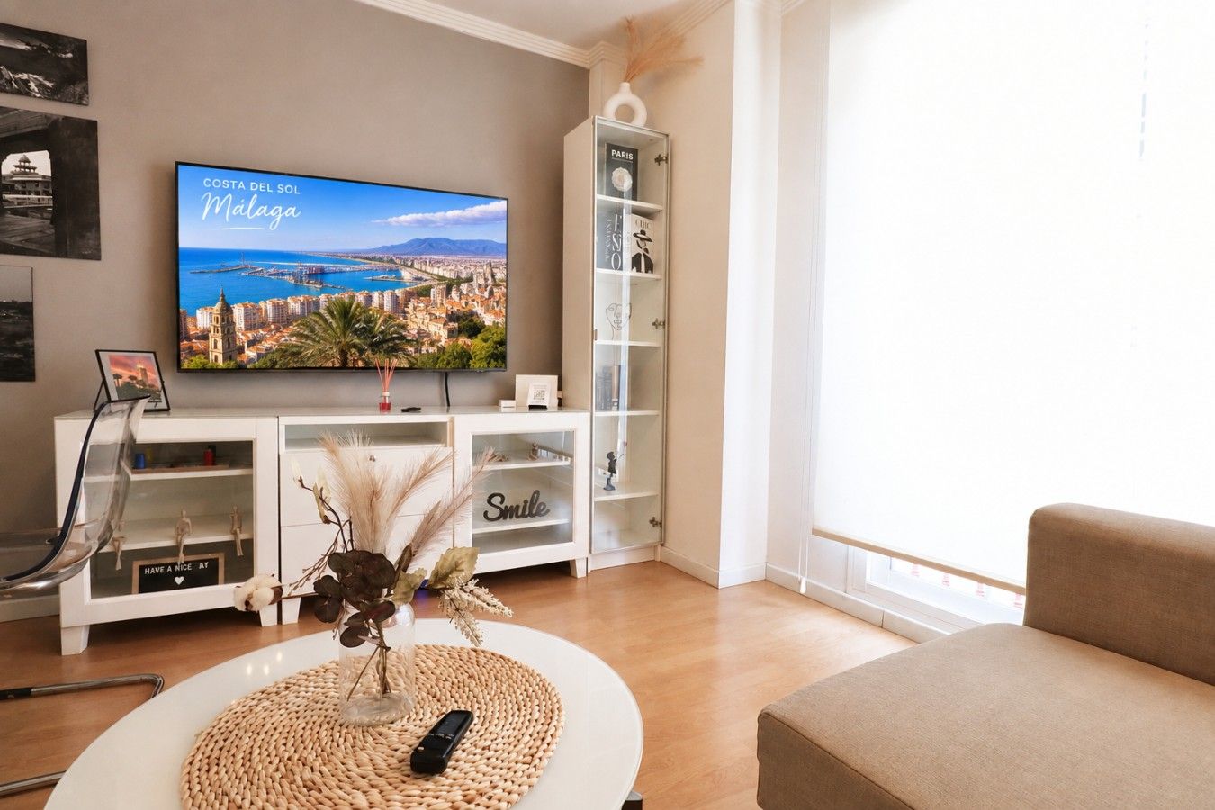 Una sala de estar con una televisión mostrando un paisaje urbano, un mueble blanco con estantes, una silla transparente y un sofá color beige.