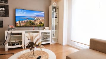 Una sala de estar con una televisión mostrando un paisaje urbano, un mueble blanco con estantes, una silla transparente y un sofá color beige.