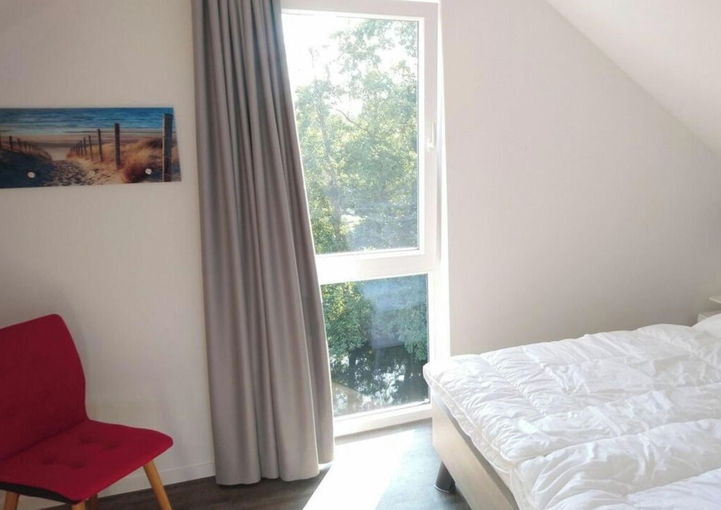 Ein Schlafzimmer mit einem Bett, einem roten Sessel, einem Gemälde und einem Fenster mit Blick auf Bäume.