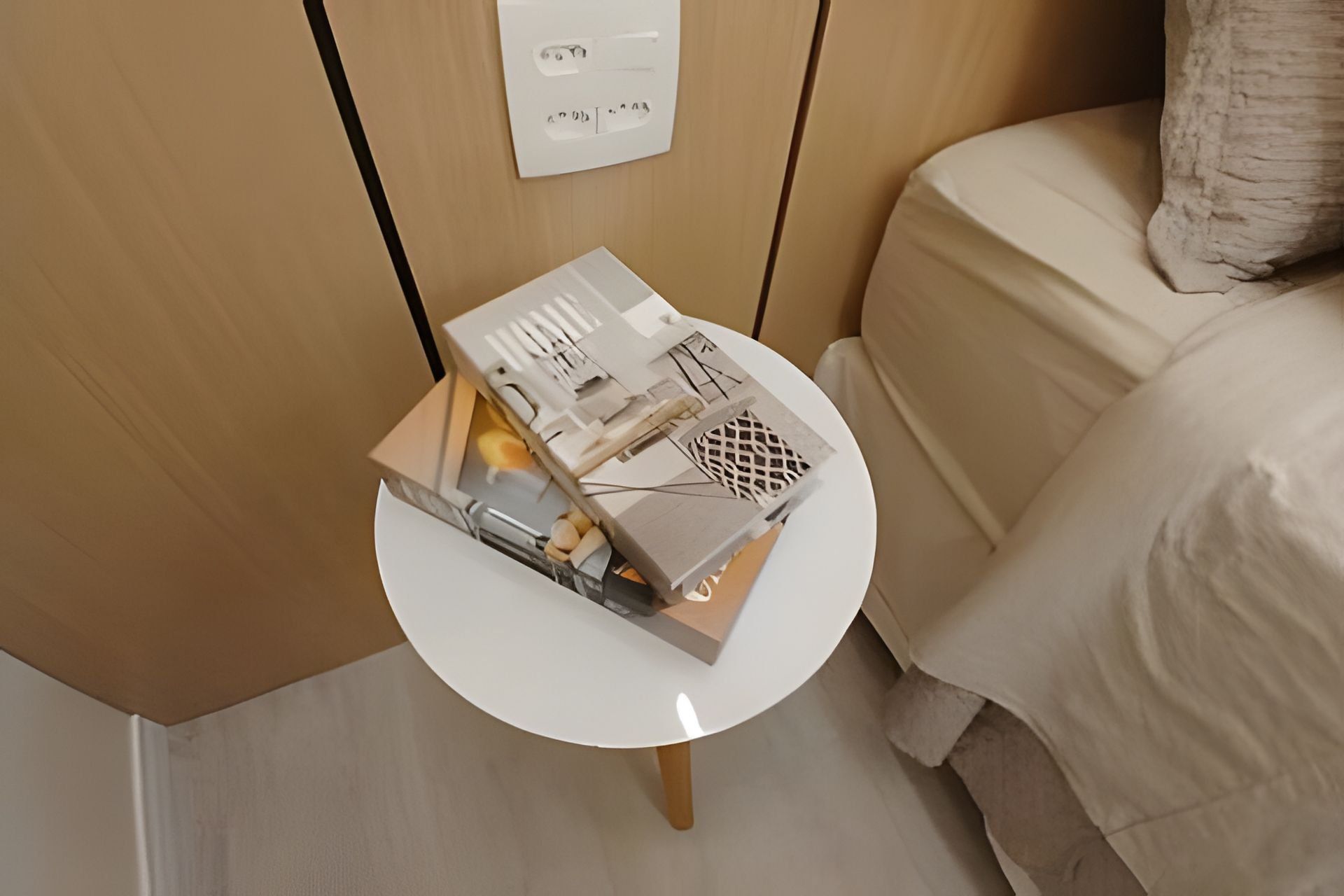 Una pequeña mesa blanca con una caja que contiene varios objetos, un enchufe y una cama con cubierta color beige.