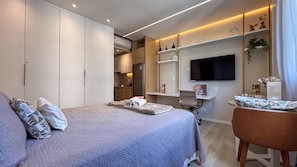 Quarto de hotel com cama, escrivaninha, televisão e cozinha compacta.