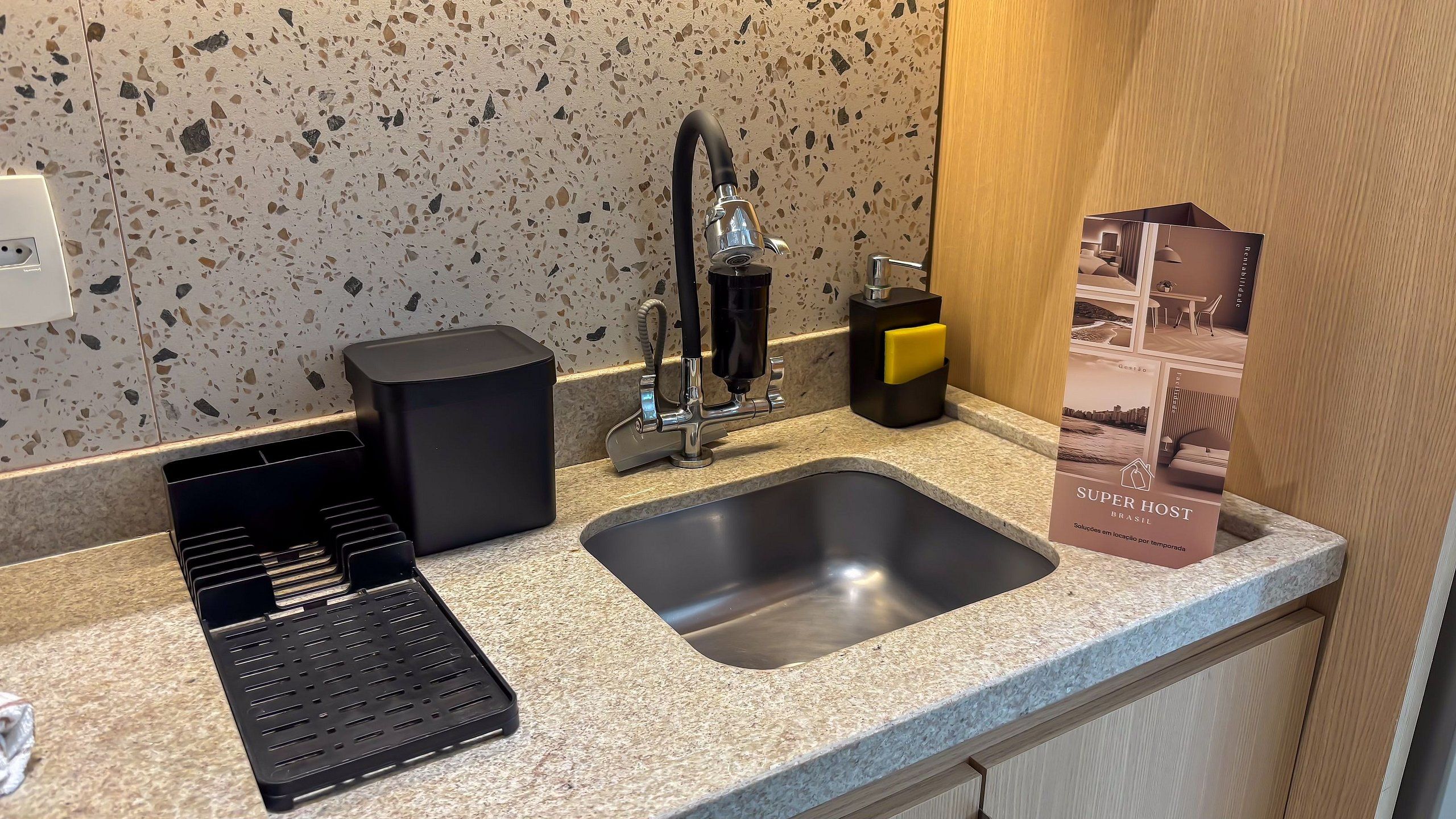 A kitchen with a granite countertop, a stainless steel sink, a black coffee machine, and a trash bin.