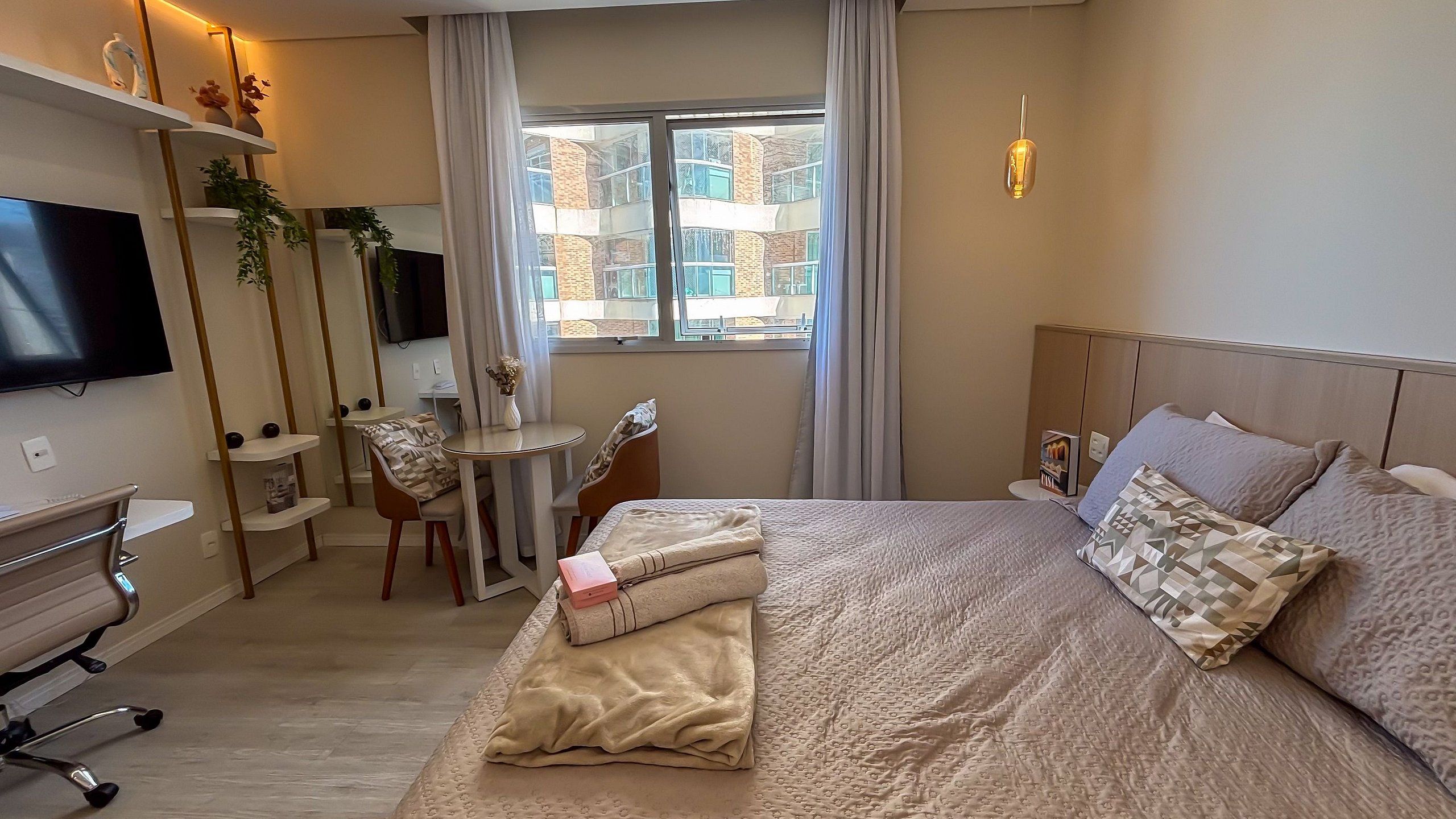 A hotel room with a bed, a desk, a chair, a TV, and a window with a view of buildings.