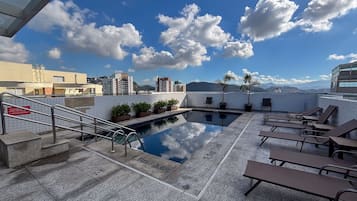 A rooftop pool with lounge chairs, a small pool area, and a view of surrounding buildings and mountains.