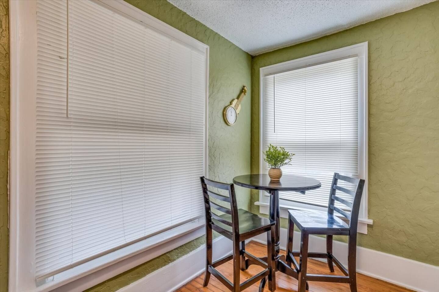 A room with two windows, a round table with chairs, and a clock on the wall.
