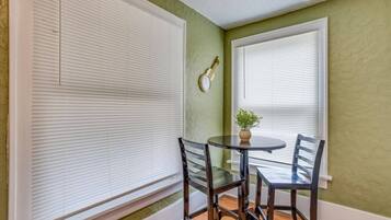 A room with two windows, a round table with chairs, and a clock on the wall.