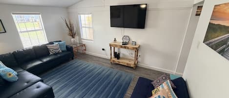 Una sala de estar con un sofá de cuero negro, un tapete azul, una mesa de centro de madera y un ventilador de techo.