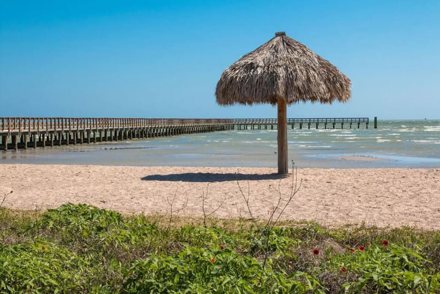 Una spiaggia con un ombrellone di paglia, un molo di legno che si protende nell'acqua e terreno sabbioso.