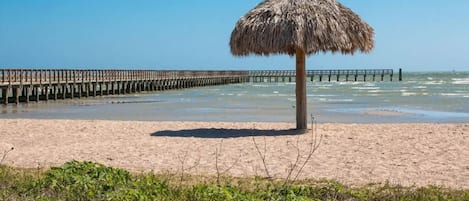 A beach with a thatched umbrella, a wooden pier extending into the water, and sandy terrain.