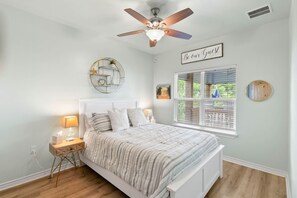 A bedroom with a bed, bedside tables, a ceiling fan, a window with blinds, and a wall-mounted sign. - Salty Vibes ~ Canal Front (Rockport)