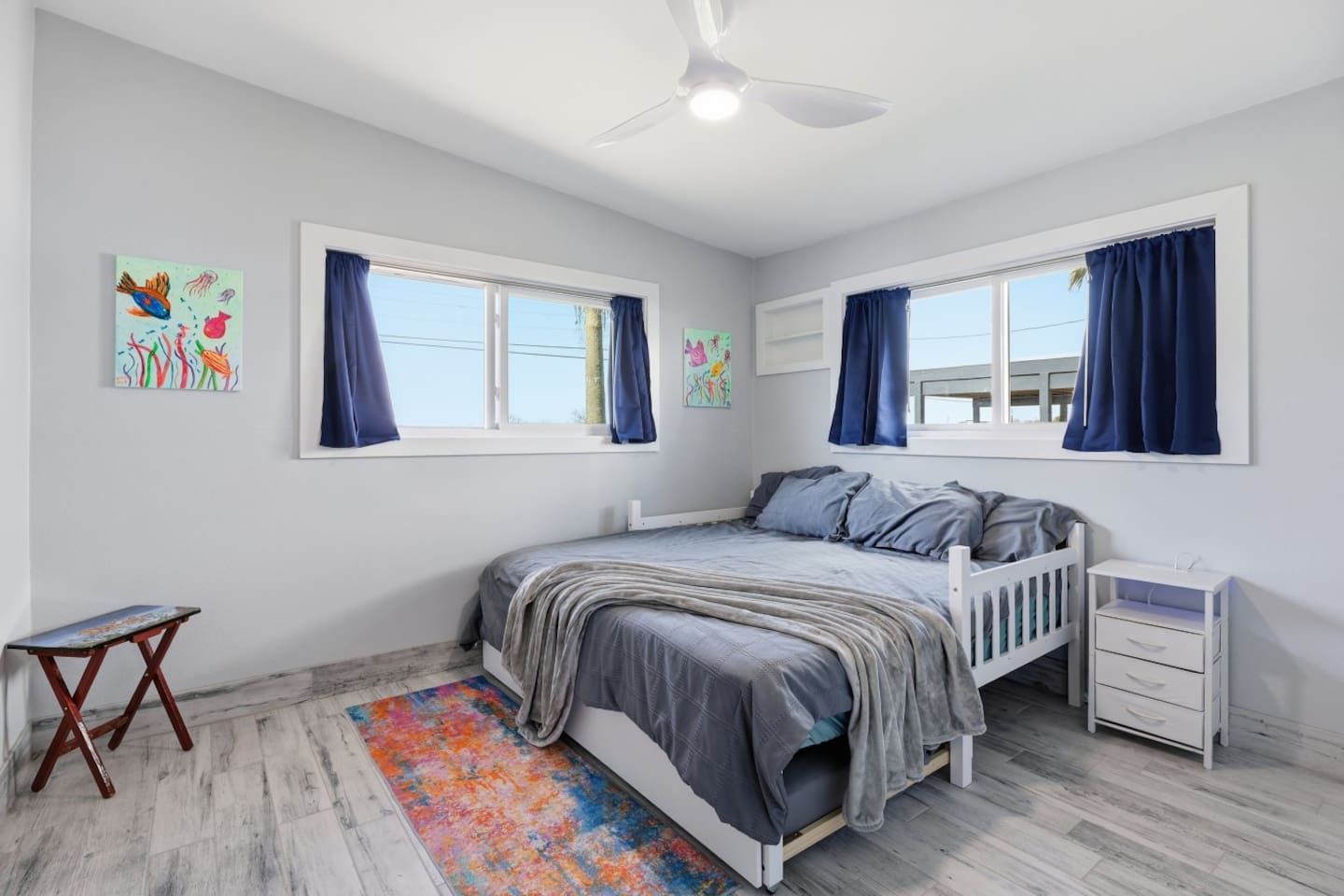 Ein Schlafzimmer mit einem Bett, zwei Fenstern, einem Deckenventilator, einem kleinen Tisch und einem farbenfrohen Teppich.