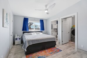 Ein Schlafzimmer mit einem Bett, einem Deckenventilator, einem Fenster mit Vorhängen, einem Nachttisch, einer Kommode und einer Waschmaschine.
