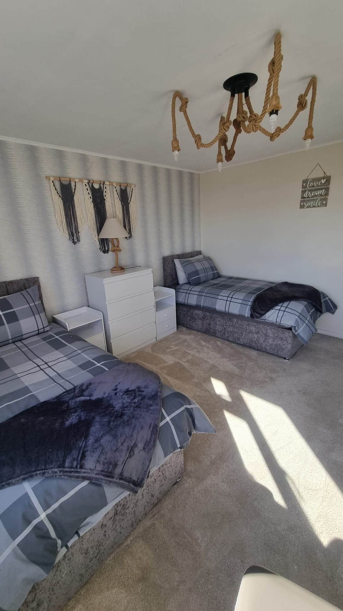 A bedroom with two beds, a white dresser, a macrame wall hanging, a chandelier, and a wall sign.