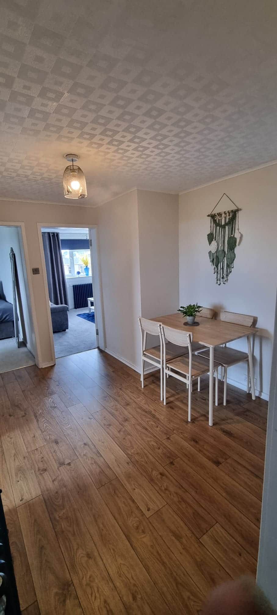 Ein Zimmer mit Holzboden, einem Tisch mit Stühlen, einer hängenden Makramee-Dekoration und einer Deckenleuchte.