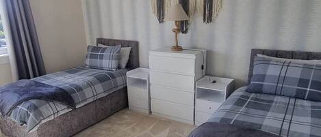 A bedroom with two beds, a chandelier, a dresser, and a macrame wall hanging.