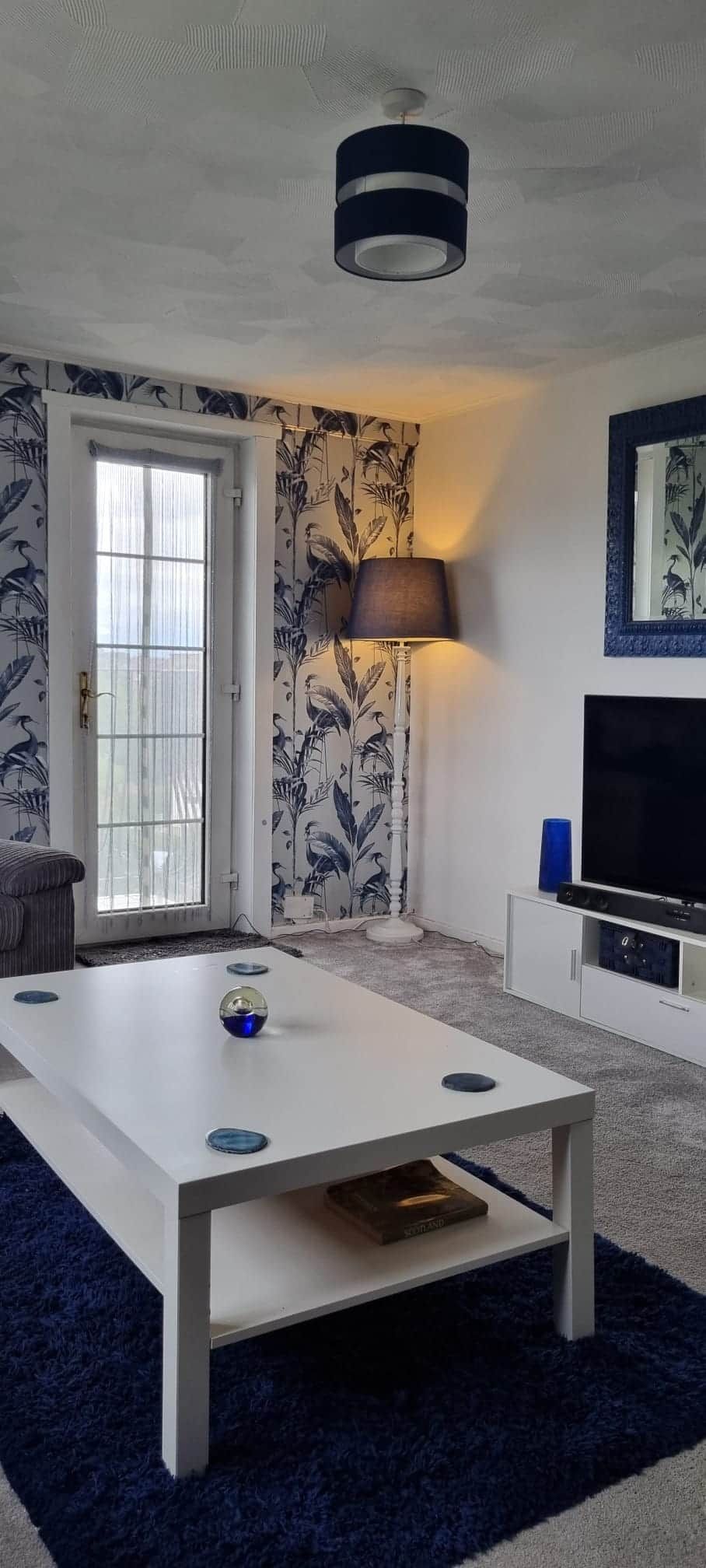 Una sala de estar con una mesa de centro blanca, una lámpara, un televisor y una puerta con ventanal.