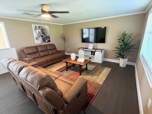 A living room with a brown leather sofa, a wooden coffee table, a television mounted on the wall, and a potted plant.