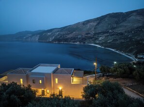 A modern two-story building with a flat roof, located near a coastline with a view of the sea and a hilly landscape in the background. - Atheras Lassu Retreat (argostoli)