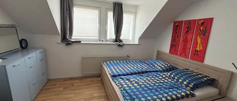 A bedroom with a sloped ceiling, a bed with a patterned bedspread, a dresser, a radiator, and a window with curtains.